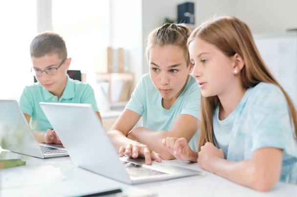 Menino e menina do ensino médio sentados ao lado da mesa em sala de aula e olhando para coisas curiosas na exibição do laptop na aula
 - Foto, Imagem