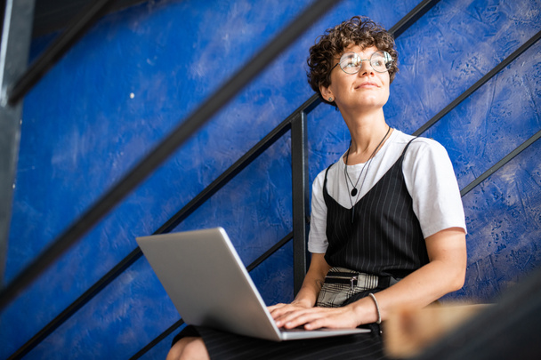 Jovem estudante moderno feliz ou gerente em casualwear usando laptop enquanto sentado na escada
 - Foto, Imagem