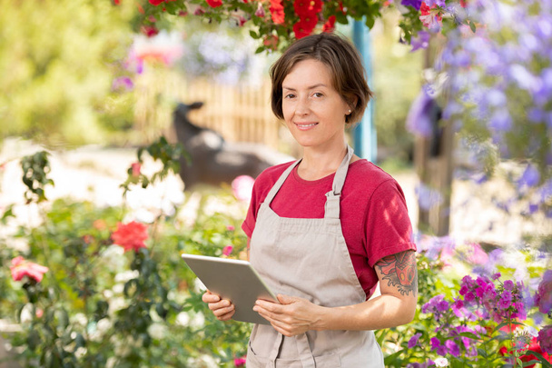 Счастливая женщина-садовница с тачпад стоя в теплице и работает над онлайн прайс-лист для растений на сайте
 - Фото, изображение