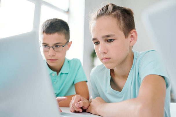 Dois meninos inteligentes sentados à mesa na frente da tela do laptop enquanto procuram dados on-line para seminário ou projeto
 - Foto, Imagem