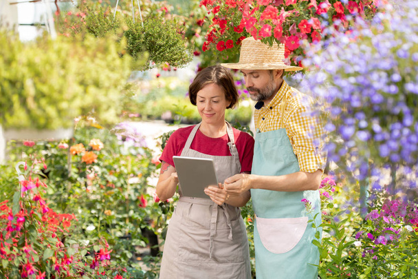 Young woman and her mature colleague in aprons looking at touchpad display while networking in greenhouse - Photo, Image