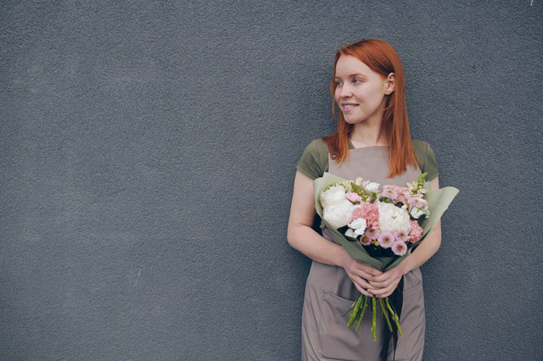 Sorrindo talentoso jovem florista do sexo feminino com cabelo vermelho vestindo avental em pé contra a parede cinza e segurando belo buquê em papel de embrulho
 - Foto, Imagem