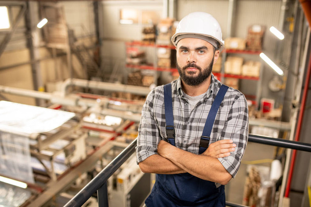 Young bearded foreman in overalls and hardhat crossing arms by chest while standing in warehouse of large factory - Photo, Image