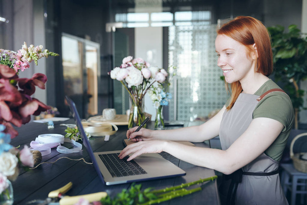Smiling attractive female florist with red hair standing at counter with flowers and packaging items and using laptop while making list of necessary flowers - Photo, Image
