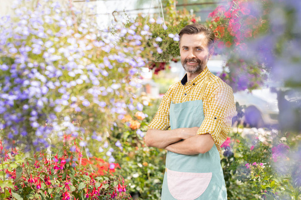 Feliz hombre jardinero cruzando sus brazos por el pecho mientras mira a la cámara dentro del invernadero lleno de flores
 - Foto, imagen