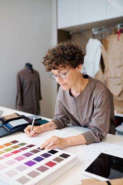 Diseñadora de moda femenina bastante joven eligiendo muestras de textiles mientras mira a través del catálogo por escritorio
 - Foto, Imagen