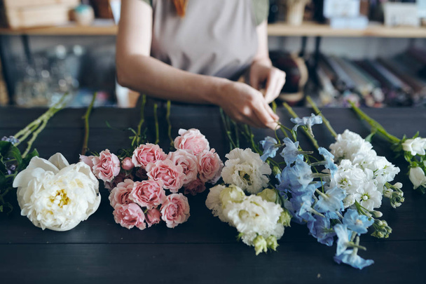 Close-up of unrecognizable florist standing at dark blue wooden table and preparing flower for arranging bouquet - Photo, Image