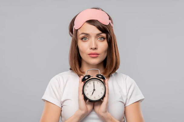 Young brunette female with alarm clock showing 7 am standing in front of camera in isolation - Photo, Image