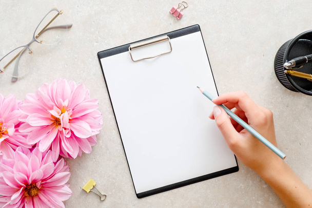 Woman hand holding pencil and writing a text into blank paper clipboard mockup. Flat lay home office desk. Top view female workspace with paper clipboard mockup, pink dahlia flowers, woman accessories - Foto, Bild