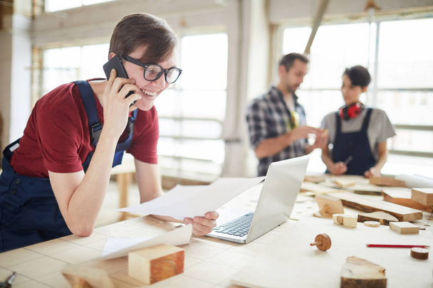 Portrait of modern carpenters working at woodworking factory, focus on smiling young man speaking by phone and using laptop in foreground, copy space - Photo, Image