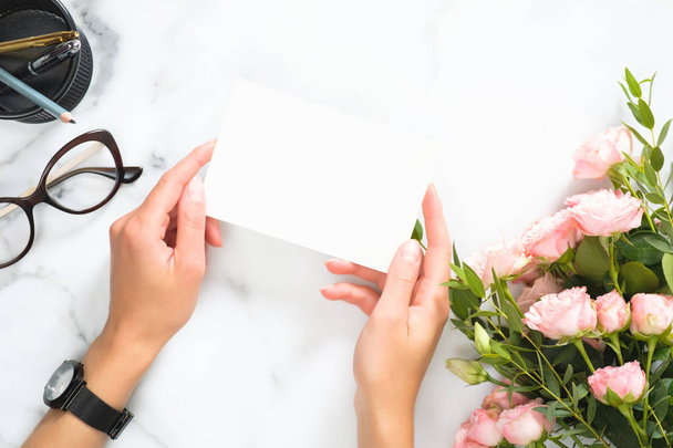 Woman hands holding blank paper card mockup over modern home office desk workspace with stationery, rose flowers bouquet, glasses, Flat lay, top view. - Φωτογραφία, εικόνα