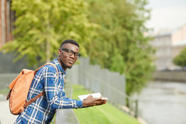 Retrato de la cintura hacia arriba del estudiante africano mirando la cámara mientras lee el libro al aire libre en la hermosa ciudad, espacio para copiar
 - Foto, Imagen