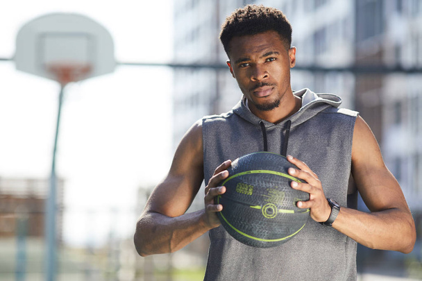 Waist up portrait of muscular African man holding basketball ball in outdoor court and looking at camera, copy space - Photo, Image