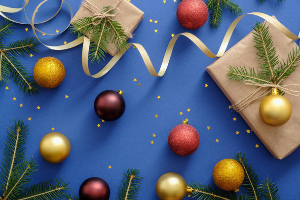 Christmas composition. Flat lay Xmas decorations, balls, presents wrapped in kraft paper, gold ribbon, fir tree branches, confetti on blue background. New Year concept or Christmas postcard mockup - Fotoğraf, Görsel