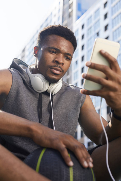 Alacsony szög arckép-ból korabeli afrikai-amerikai ember használ smartphone rövid idő ülő-ban kosárlabda udvar, másol hely - Fotó, kép
