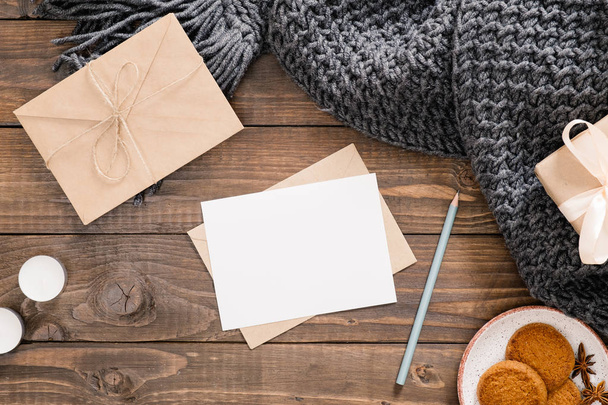 Cozy home desk, hygge, autumn fall concept. Blank paper card mockup, craft envelope, cookies, candles, women fashion scarf, gift box on wooden background. Flat lat, top view, copy space. - Φωτογραφία, εικόνα