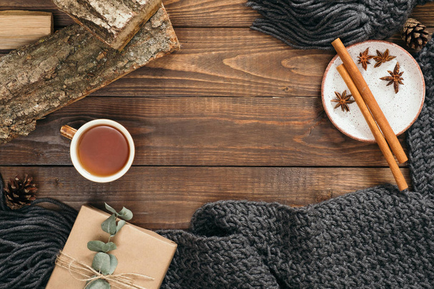 Flatlay hygge style composition with fashion women knitted scarf, mug of tea, gift box, firewood, cinnamon sticks on wooden background. Flat lay, top view, overhead. Cozy autumn home desk concept - Фото, изображение