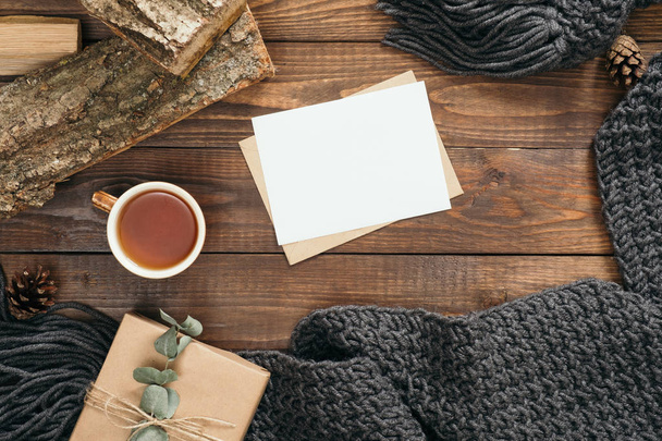 Flatlay hygge style composition with fashion women knitted scarf, letter, blank paper card, mug of tea, present, firewood on wooden background. Flat lay, top view, overhead. Cozy autumn home desk - 写真・画像