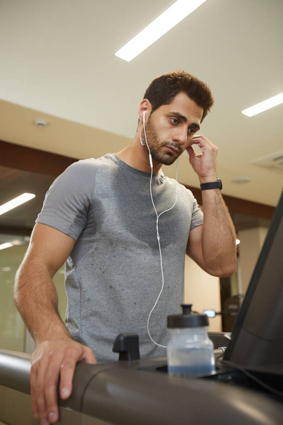 Taille hoch Porträt eines verschwitzten jungen Mannes, der Musik hört, während er im Fitnessstudio auf dem Laufband läuft - Foto, Bild