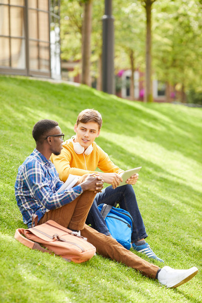 Retrato de vista lateral de dos estudiantes sentados en césped verde en el campus y relajantes al aire libre, espacio para copiar
 - Foto, Imagen
