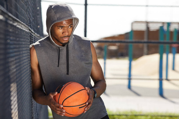 Μέση επάνω πορτρέτο του σύγχρονου Αφροαμερικανικού ανθρώπου κρατώντας μπάλα μπάσκετ που ποζάρει σε αθλητικό γήπεδο σε εξωτερικούς χώρους, αντιγραφή χώρου - Φωτογραφία, εικόνα