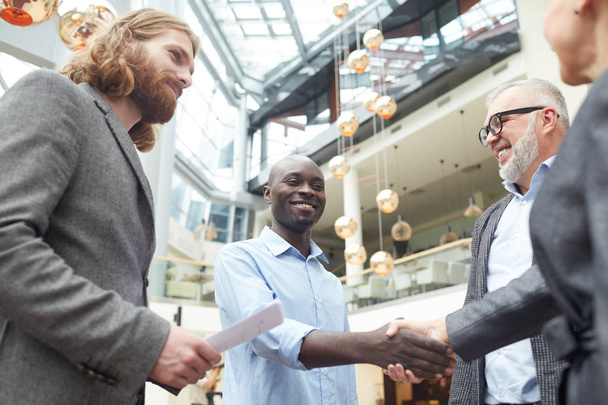 Retrato de baixo ângulo de grupo de empresários apertando as mãos com o homem afro-americano de pé no salão de escritório após entrevista de emprego bem sucedida, espaço de cópia
 - Foto, Imagem