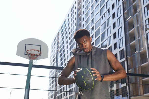 Cintura-se retrato de homem afro-americano bonito segurando bola de basquete e olhando para a câmera com confiança enquanto está em pé na quadra ao ar livre, espaço de cópia
 - Foto, Imagem