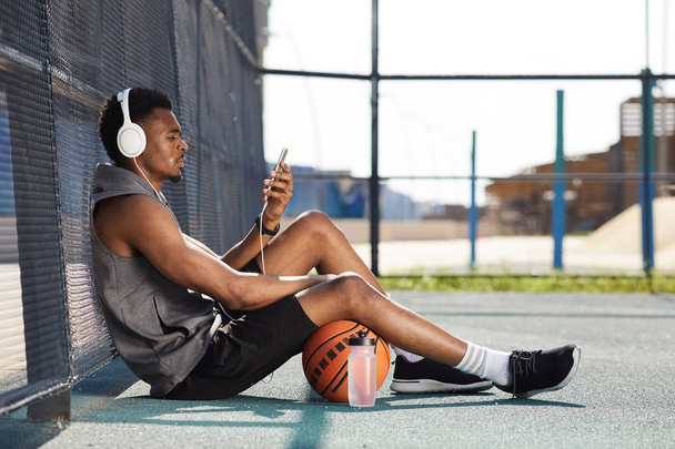 Πλευρική όψη πορτραίτο του σύγχρονου Αφρο-Αμερικάνικου άνδρα που φοράει ακουστικά και χρησιμοποιεί smartphone ενώ κάθεται σε γήπεδο μπάσκετ σε εξωτερικούς χώρους, αντίγραφο χώρου - Φωτογραφία, εικόνα