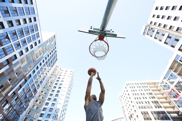 Χαμηλή γωνία δράσης σφαίρα του Αφρικανός παίκτης μπάσκετ γυρίσματα χτύπημα ενάντια στον ουρανό σε αστικό φόντο, αντίγραφο χώρου - Φωτογραφία, εικόνα
