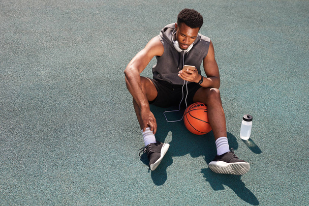 Acima de fundo vista de homem afro-americano bonito sentado na quadra de basquete e usando smartphone, espaço de cópia
 - Foto, Imagem