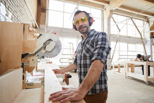 Waist up portrait of mature carpenter wearing protective gear smiling at camera while sawing wood in industrial workshop, copy space - Photo, Image