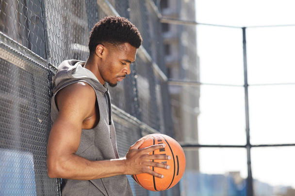 屋外のスポーツコートでフェンスに立ちながらバスケットボールボールを持つ筋肉質のアフリカ系アメリカ人男性のサイドビューの肖像画、コピースペース - 写真・画像