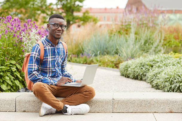 Volledige lengte portret van Afrikaanse student glimlachend op de camera tijdens het gebruik van laptop buitenshuis in College campus, kopieer ruimte - Foto, afbeelding
