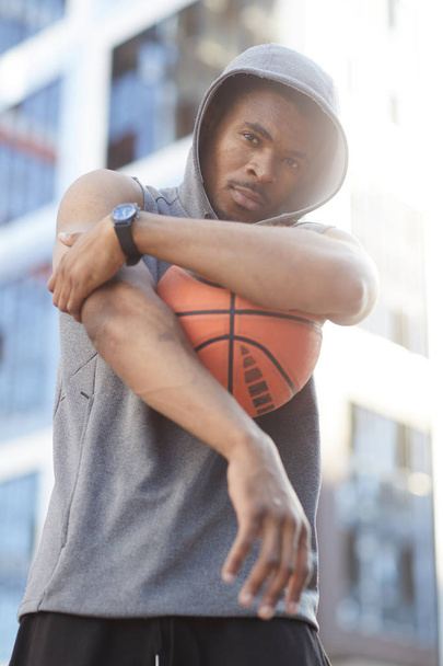 Waist up portrait of contemporary African man wearing hoodie posing with basketball ball in urban setting - Zdjęcie, obraz