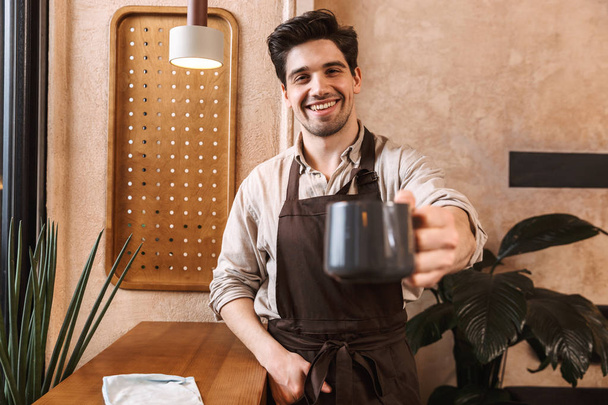 Cheerful men barista wearing apron standing at the cafe - Фото, изображение