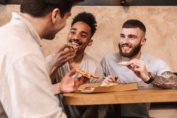 Three cheerful men eating pizza at the cafe table - Fotó, kép