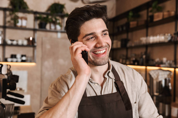 Handsome happy coffee man posing in cafe bar working indoors talking by mobile phone. - Fotó, kép