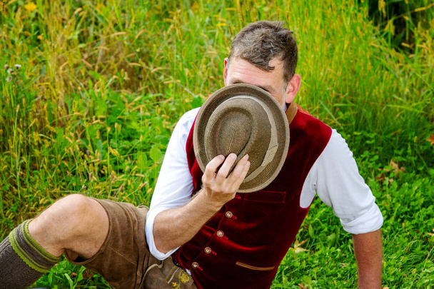 bavarian man sitting on grass and hiding behind his hat - 写真・画像