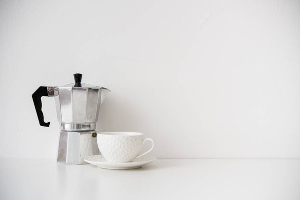Metalli kahvinkeitin ja valkoinen posliini kuppi pöydälle tyhjä seinä
 - Valokuva, kuva