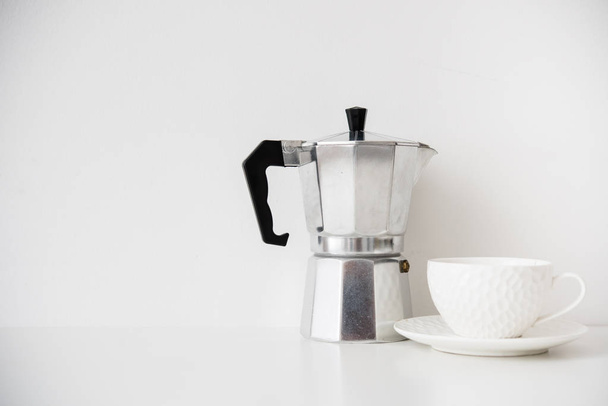 Fém kávéfőző és fehér porcelán csésze az asztalon, üres fallal - Fotó, kép