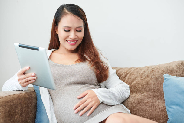 Mosolygó fiatal ázsiai terhes nő pihenő a kanapén tabletta számítógép kezében, és megérintette a hasát - Fotó, kép