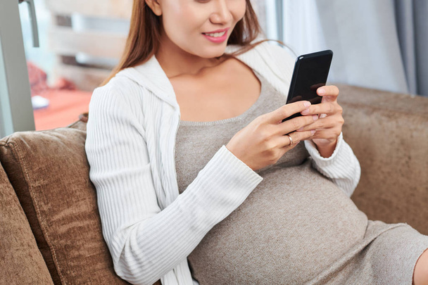 Cropped image of positive young pregnant woman sitting on comfy sofa at home and checking social media on her smartphone - Photo, Image