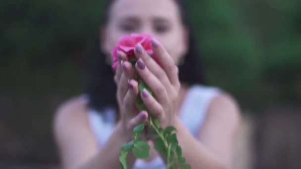 Adult beautiful brunette lady in white dress smells flower in the garden. Woman extends a hand with a flower to the camera. Time pending outdoor at green garden. Female is smiling and happy - Metraje, vídeo