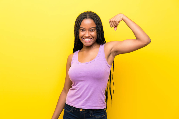 afrikanisch-amerikanisches Teenager-Mädchen mit langen geflochtenen Haaren über isolierter gelber Wand mit starker Geste - Foto, Bild