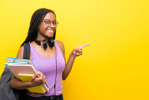 afro-américaine adolescent étudiant fille avec de longs cheveux tressés sur isolé mur jaune pointant doigt sur le côté
 - Photo, image