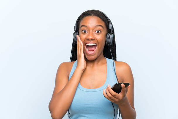 Afro-amerikai tinédzser lány hosszú fonott hajú hallgató zene egy mobil a meglepetés és sokkolta arckifejezés - Fotó, kép