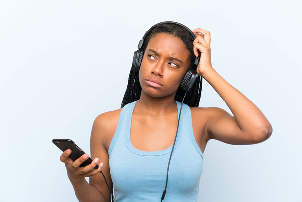 Menina adolescente afro-americana com cabelos longos trançados ouvindo música com um celular com dúvidas e com expressão facial confusa
 - Foto, Imagem