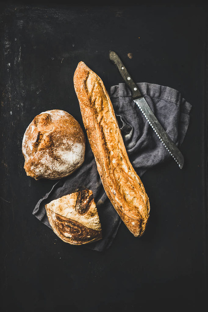 Flat-lay of freshly baked baguette and loaf on dark linen over rustic black background, top view - Фото, зображення