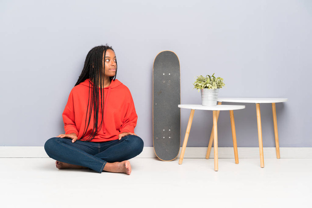 African American skater teenager girl with braided hair sitting on the floor looking to the side - Foto, Bild