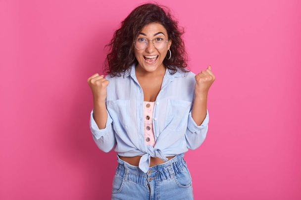 Image of adorable brunette woman with dark swavy hair, clenches fists and exclaims with triumph expression, screaming; Yes. Attractive female wearing shirt and jeans. People and body language concept. - Foto, afbeelding